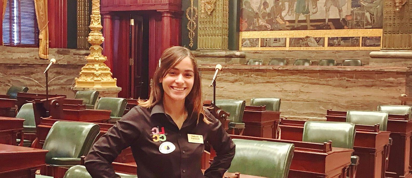 A young woman stands with her hands in her hips in the middle of a large congressional assembly room.